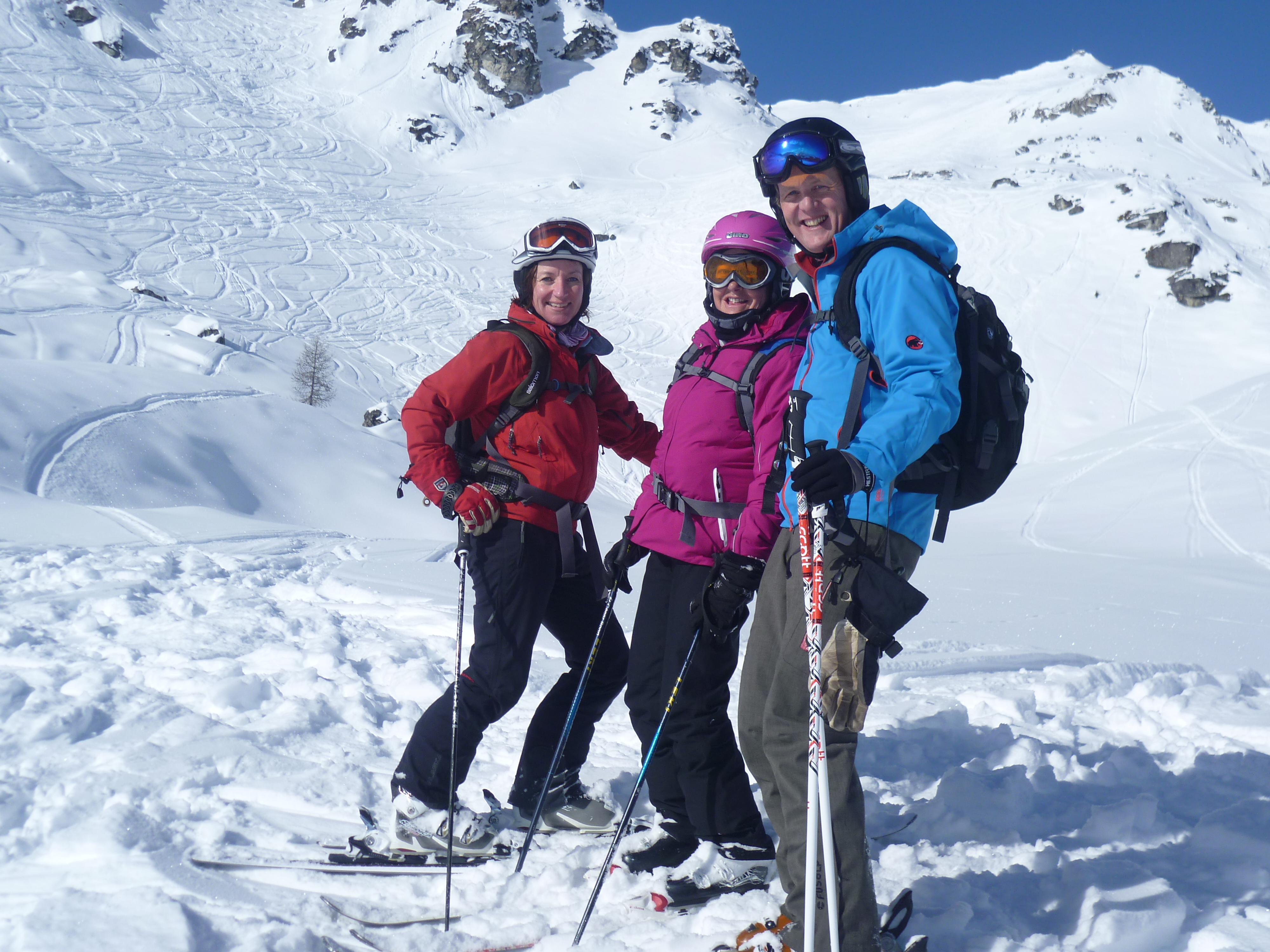 Three skiers posing on a snowy slope, wearing colorful ski gear with mountains in the background.