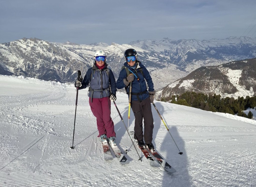 Two skiers wearing helmets and goggles stand together on a snowy mountain slope, with a scenic backdrop of snow-covered peaks and valleys.