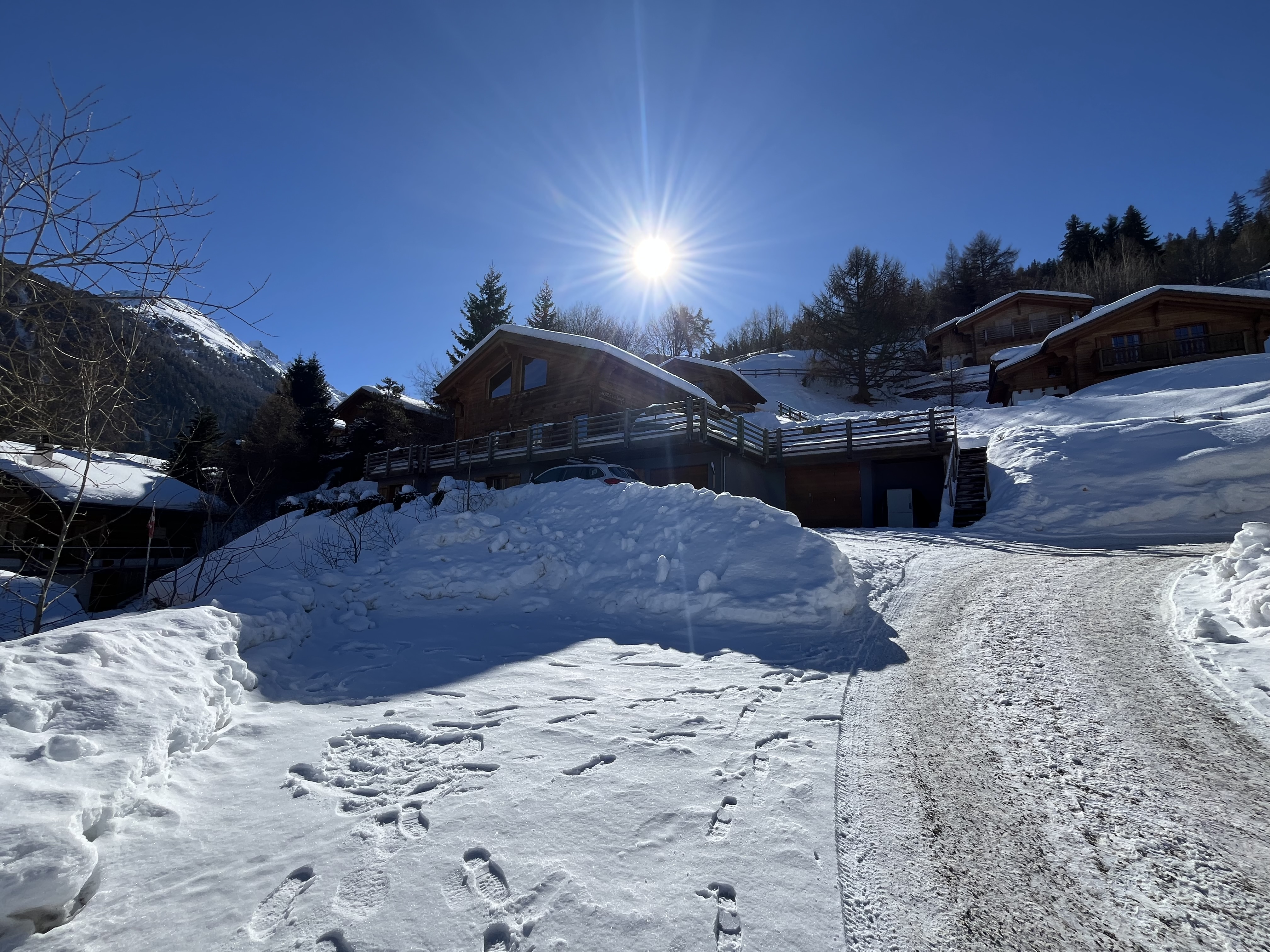 A sunny winter scene featuring wooden chalets surrounded by snow-covered ground and mountains in the background.