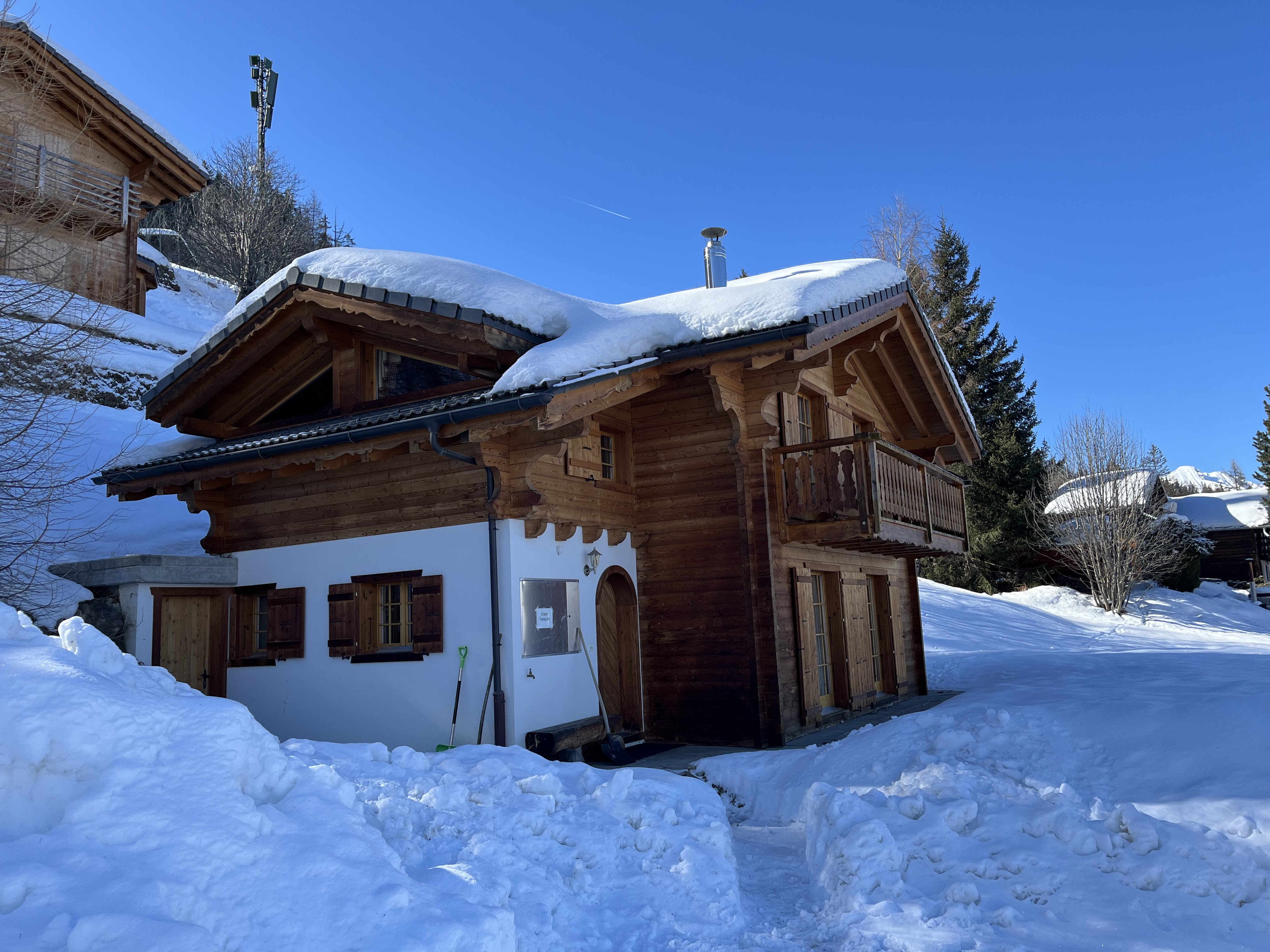 A wooden chalet covered in snow, with a steep roof and a balcony, set against a clear blue sky.