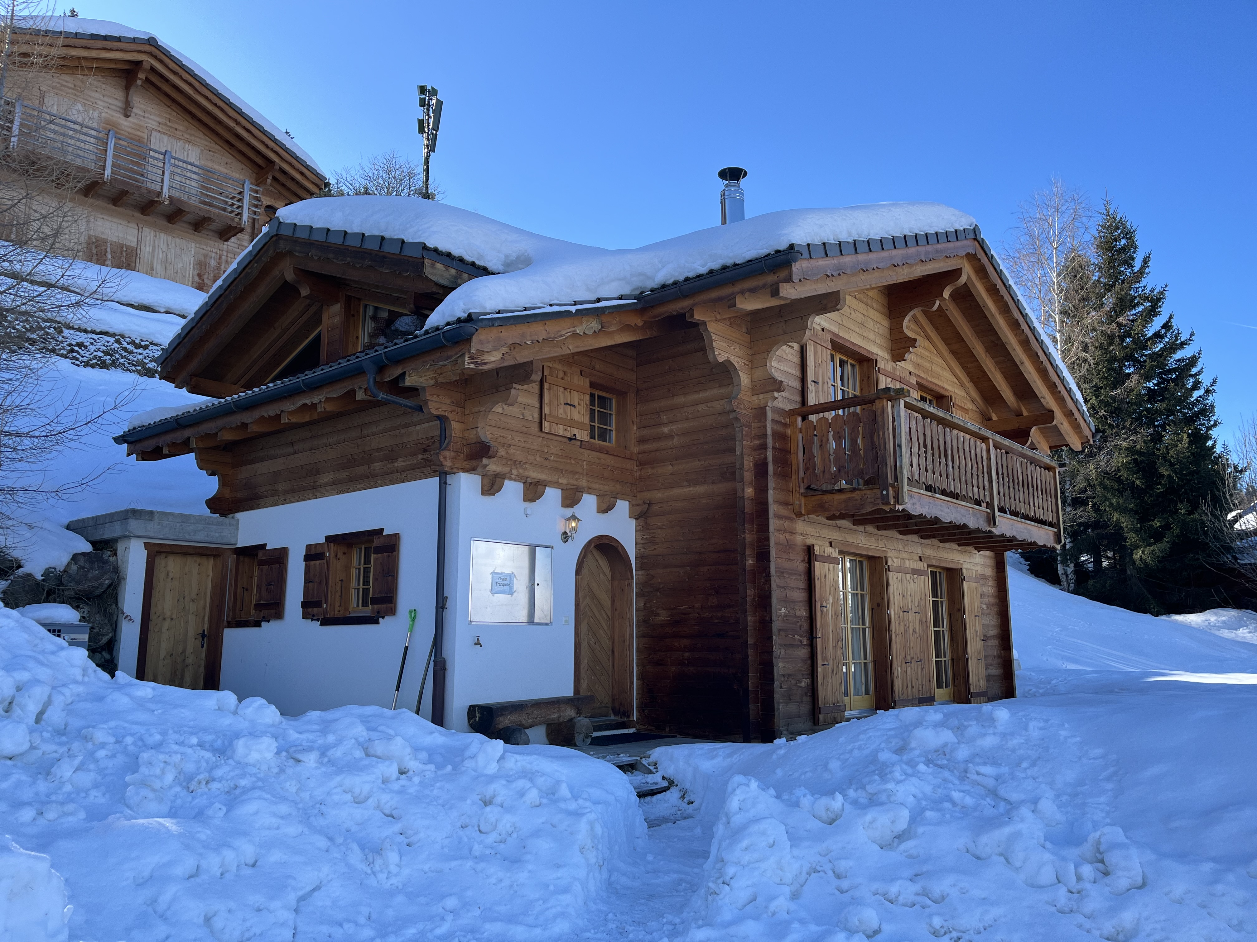 A charming wooden chalet with a sloped roof covered in snow, situated in a winter landscape. The chalet features a balcony, large windows with wooden shutters, and a snow-covered path leading to the entrance.