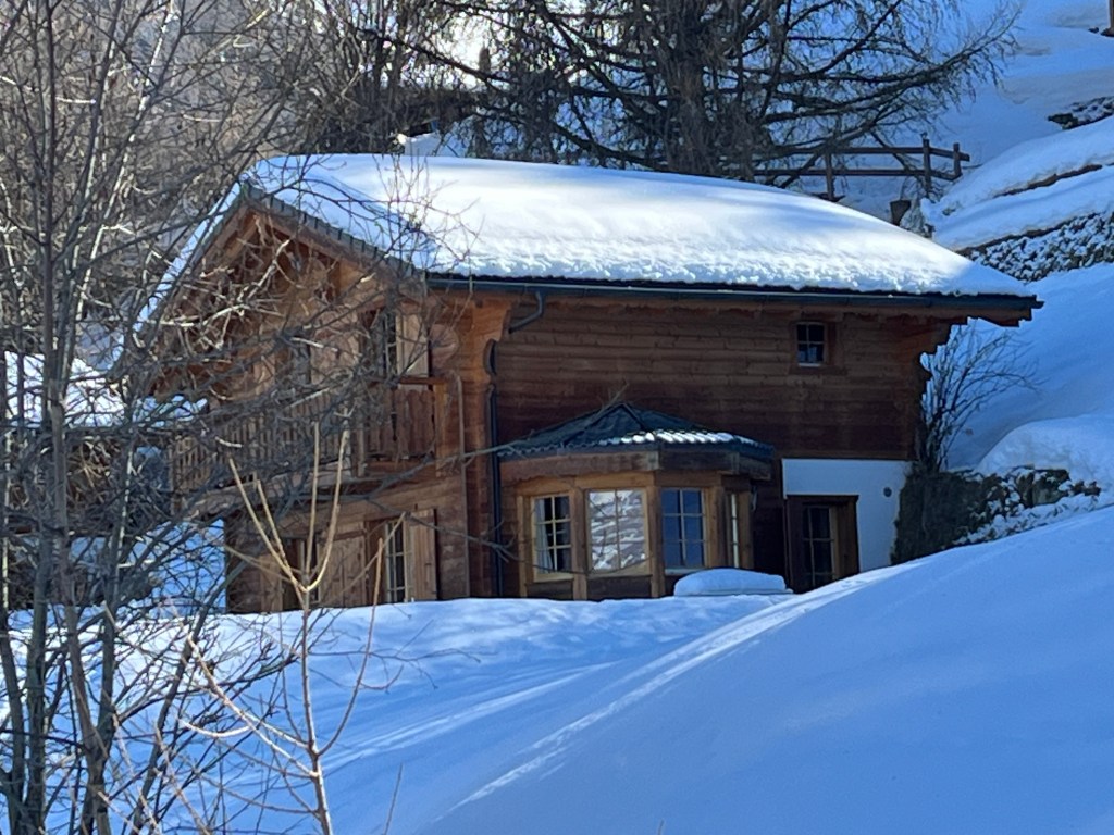 A wooden chalet with a sloped roof covered in snow, surrounded by trees and rolling white hills.