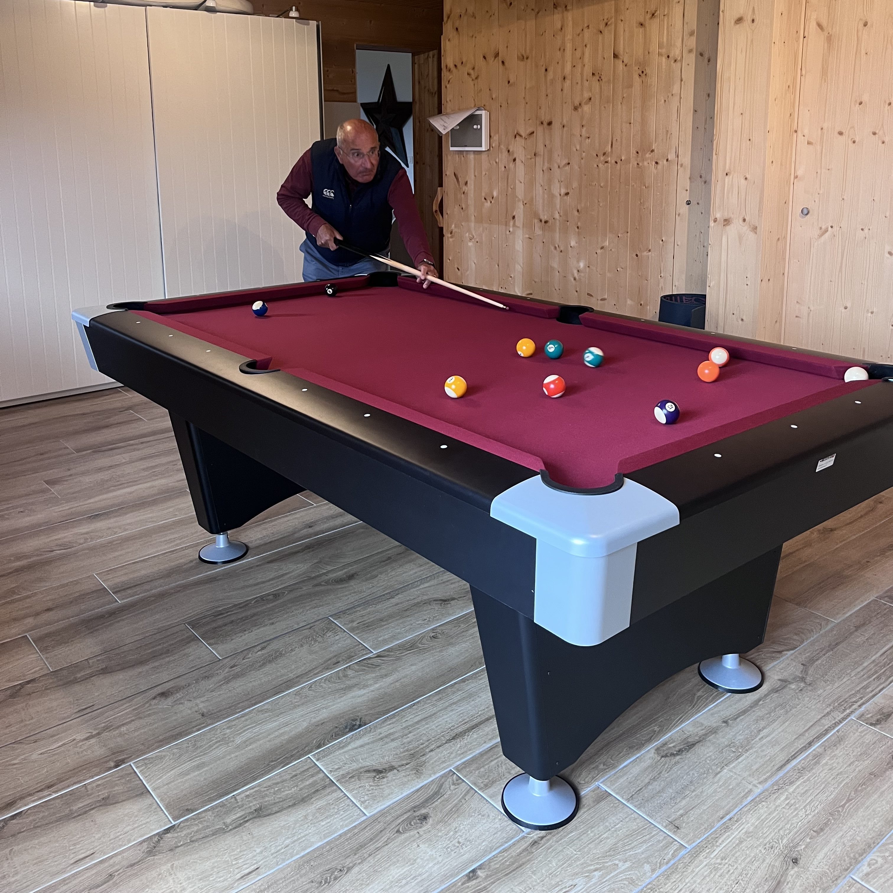An individual is playing pool on a professional billiard table with a maroon felt surface. The player is focused on a shot, with various colored balls positioned on the table in a well-lit wooden room.
