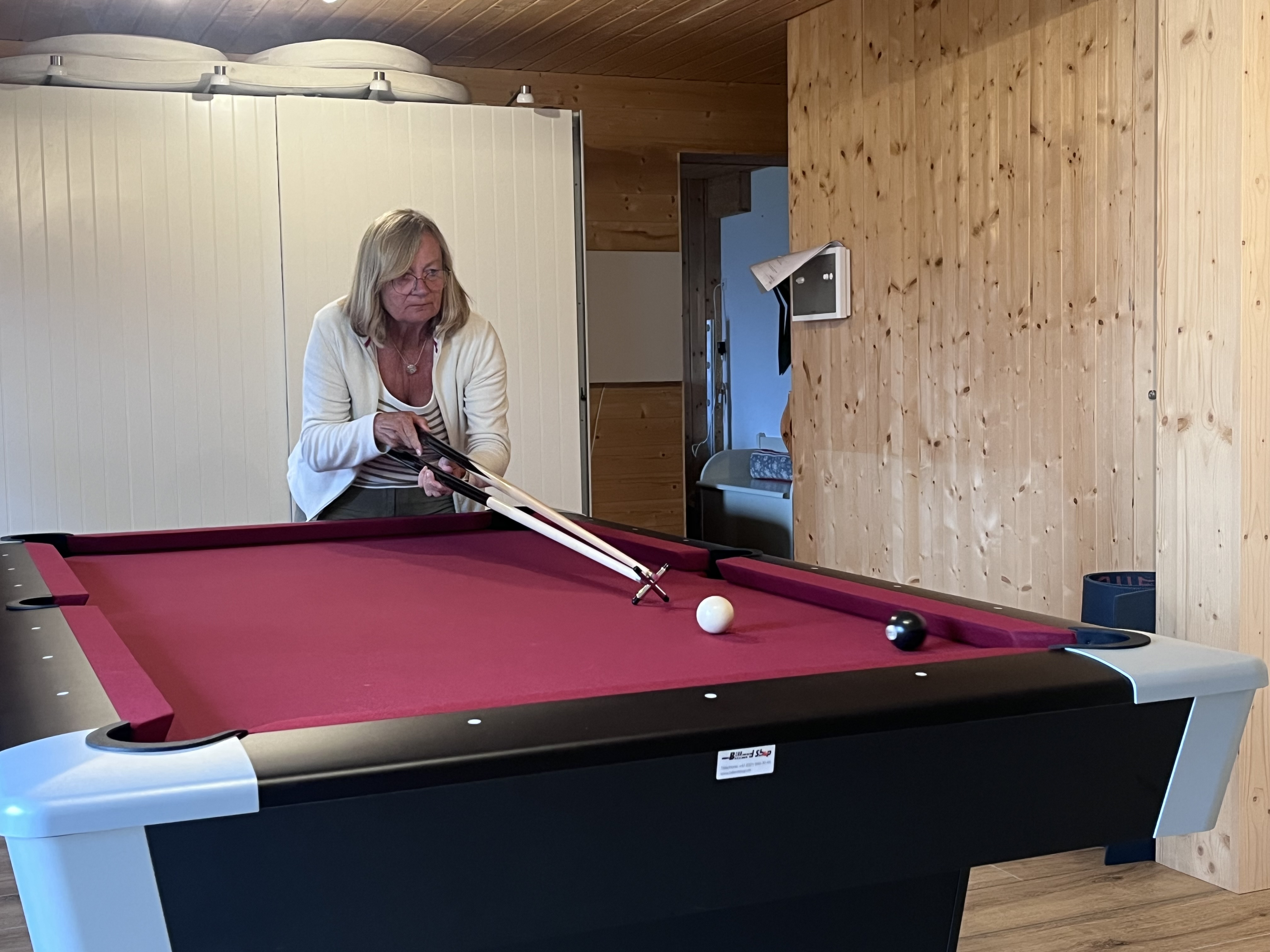 A woman with long hair and glasses is preparing to take a shot at a billiard table with a pink felt surface, holding two cues. The background features wooden walls and a glimpse of a room.
